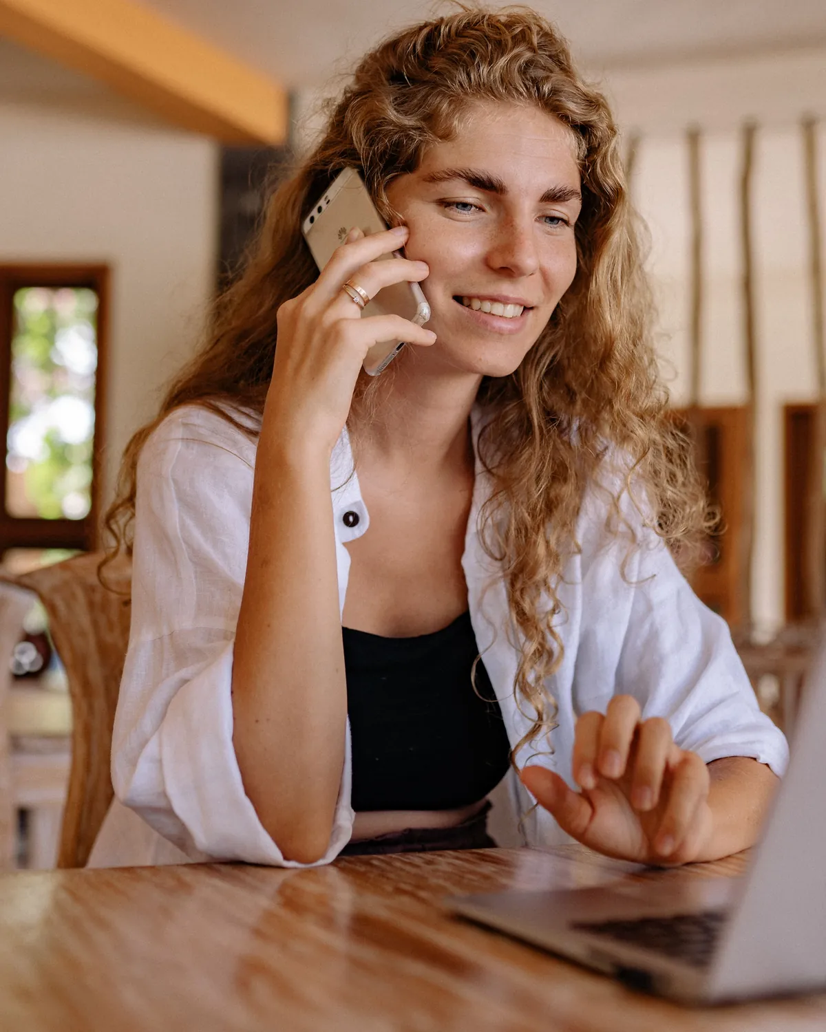 Femme souriante au téléphone, assise à une table avec son ordinateur portable, dans un intérieur chaleureux